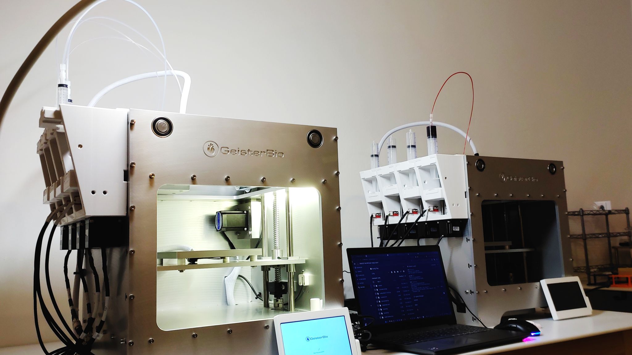 Two GeisterBio MAK007-4X bioprinters on a lab bench, connected to syringe extruders, touchscreen controls and a laptop running the control software