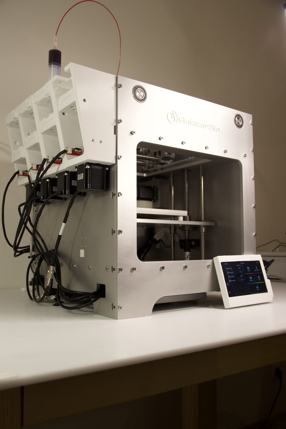 GeisterBio MAK007-4X bioprinter on a bench, showing the four-slot multi-material printhead carriage, stepper drives and a touchscreen running the control software